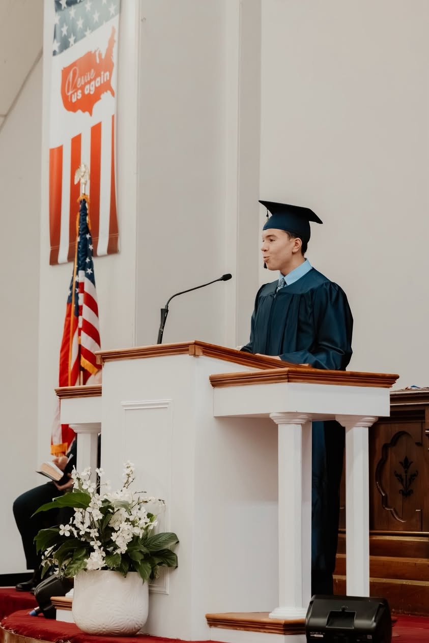 A NECA graduate in cap and gown speaking from the pulpit at graduation.